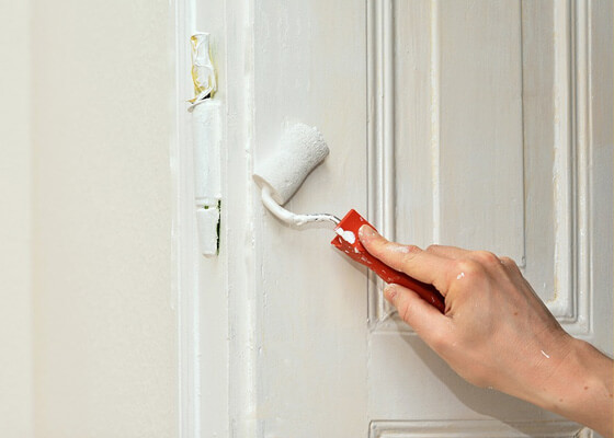 A hand holding a red-handled brush is painting a wooden door white. The door has detailed paneling, and the brush is applying fresh paint smoothly, covering the surface evenly. The background is a light-colored wall.