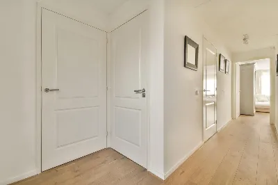 A bright, minimalist hallway with light wooden flooring. Two white doors are on the left, and framed pictures adorn the right wall. At the end of the hallway, sunlight streams into a bedroom.